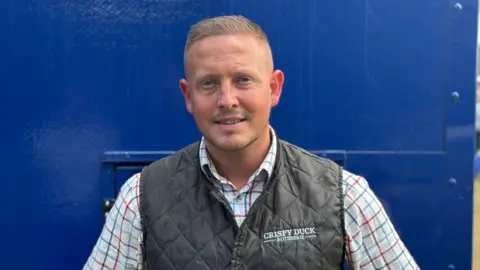 George King/BBC A head and shoulders shot of Shaun Sabiny, who is standing in front of a navy blur food trailer. He is wearing a gilet over a shirt and is smiling into the camera.