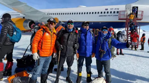 Hari Budha Magar Hari Budha Magar and three of his climbing team arriving in Antarctica. Hari, pictured second from left, is wearing a cap, sunglasses, puffer jacket and shorts. He is holding two walking sticks. An aeroplane with passengers disembarking can be seen in the background.