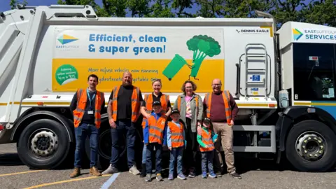 East Suffolk Services Five adults and three children in front of them stand in front of a bin lorry. All are wearing orange hi-vis tabards and smiling at the camera. 