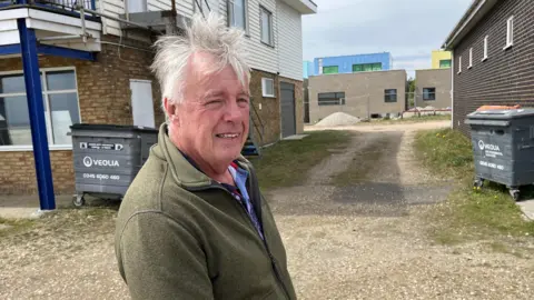 Phil Harrison/BBC Tony Malthouse, wearing a green jumper. Blue and yellow coloured houses can be seen down a gravel path behind him.