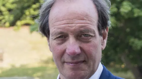 Norfolk County Council Andrew Jamieson looking directly at the camera. He is outside on a sunny day with foliage in the background.