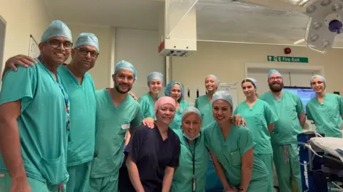 South Tyneside and Sunderland NHS Foundation Trust A team of 12 medical professionals in a theatre dressed in green scrubs. They have their arms around each other and are smiling at the camera.