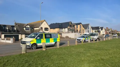 Andrew Turner/BBC An ambulance incident command unit and police car are parked on Marine Parade in Gorleston. In the foreground is a grassed area, with wooden posts providing a boundary to the pavement. In the backdrop are several houses.