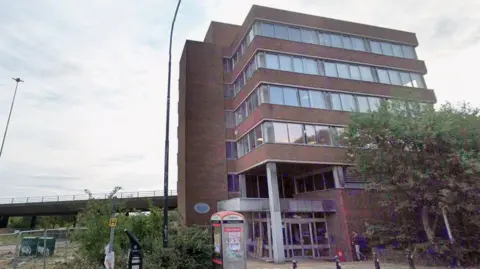 Google Computer House before its demolition. It is a nondescript, square six-storey brick office block with a glazed entrance in one corner. There is a telephone box outside, covered in the remnants of posters and graffiti. Waste ground sits to the left of it and the flyover can be seen in the background.