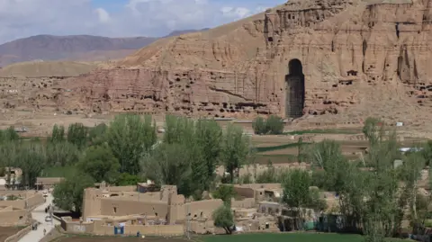 EPA/REX/Shutterstock Large hole in hillside in Bamiyan, Afghanistan, where a giant Buddha statue once stood, pictured in 2024. The hole is within cliffs of bare, red-tinged stone, there are trees and low-rise buildings in the foreground.