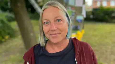 George Carden/BBC A woman with short grey/blonde/blue hair looks at the camera. She is wearing a brown hoodie and a black T-shirt. 