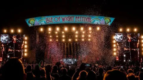 Isle of Wight Festival The main stage at the Isle of Wight festival at night time. Confetti is in the air over the heads of crowds of people who are watching the stage. A musician can be seen on the stage holding at guitar.