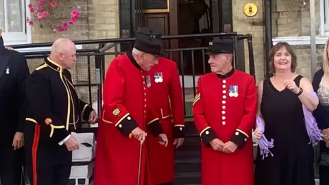 A close-up of group of men and women standing outside Kitchener's Guest House. In the middle are three male Chelsea Pensioners, clad in bright red, with a man in a formal black regimental suit on the left and a woman in a black evening dress on the right.