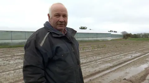 Didier Helio stood in a waterlogged field. He's wearing a black rain mac. He has short grey hair. Poly tunnels are visible in the background. 