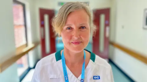 Nikki Fox/BBC Sara Heritage stands in the corridor at the West Norwich Community Hospital in Norwich. There are red doors behind her, She is wearing white scrubs with a dark green trim and has a blue lanyard around her neck. You can see the NHS logo on her top. She has blonde hair tied back and is looking at the camera. Her skin is freckled.