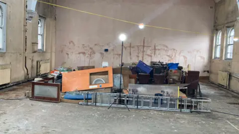 A stack of things including a door and a set of ladders, piled up in the middle of an otherwise empty room. There is a light attached to a wire strung across the centre of the room. The walls appear to have been stripped ready for decoration.