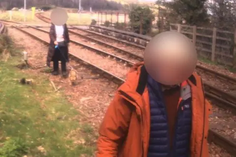Network Rail Two people, their faces blobbed out, walk along a railway line. The person nearest the camera is wearing a thick red jacket over a padded jacket. There is a person behind them wearing a long jacket and walking a small brown dog.