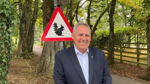 Constable Andy Jehan stood on one of the green lanes wearing a navy suit jacket and white shirt. There is a concrete path, trees and a road sign with a squirrel on it in the background.