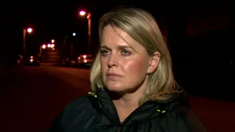 Clare Loughran speaks to a BBC reporter on a dimly lit street. She has shoulder-length blonde hair and is wearing a dark raincoat.