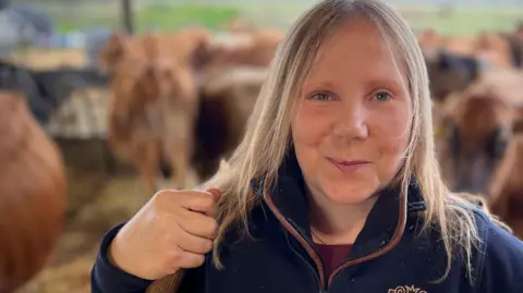 Rebecca Mayhew, a woman who is standing among cattle at a dairy farm. She is looking directly at the camera and smiling and is wearing a blue fleece jacket which says OLD HALL FARM on it.