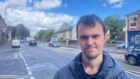 A man stands in front of a busy road with houses and shops. He has brown hair, a beard and is wearing a blue coat with a hood.