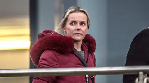 Emma Gardner, a blonde woman with her hair tied back. She has a red coat on with a red fur hood. 