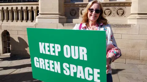 Nadia Lincoln/LDRS A woman in a white top wearing sunglasses stands in front of a stone building holding a green sign with "Keep our green space" written on it. 