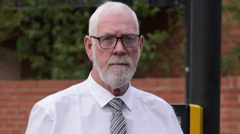 A close-up of a Gerry Duffy seen with short white hair, a neat white beard and moustache, and black-rimmed glasses looking directly at the camera with a somber expression. He is wearing a formal white collared shirt and a diagonal grey and white striped tie. The background shows a blurred outdoor setting with a red brick wall, green foliage, and a dark pole.