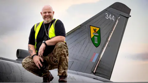 Andrew Wheeler. He appears to be posing on the wing of a fighter jet. He is wearing khaki trousers, a black t-shirt and a hi-vis vest.