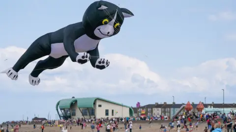 A huge black and white cat kite can be seen flying over the beach on Saturday. It can be seen over people walking on the beach and a cream building in the background.
