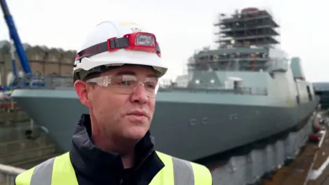 A man in a florescent jacket wearing a white hard hat with goggles is standing in front of a large grey warship.