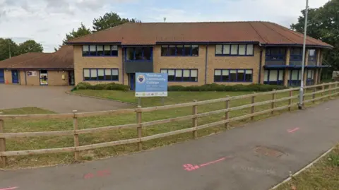 An image of Thurston Community College. It is a brick building with blue trim around the windows. A sign in front of the building says Thurston Community College. A path runs in front of a wooden fence. 