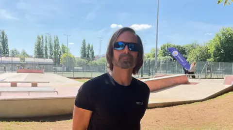 Russ Holbert wearing a black T-shirt and sunglasses in front of the skatepark