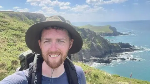Handout Aidan Courts taking a selfie high up near cliffs overlooking the sea. The shoreline can be seen behind him with rocks around the beach. He is wearing a hat and a rucksack and a blue T-shirt.