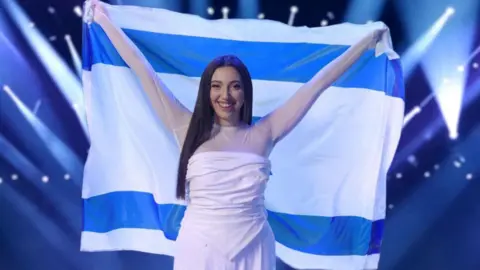 Yuval Raphael on stage holding an Israeli flag. She has long straight brown hair and is wearing a light-coloured dress.