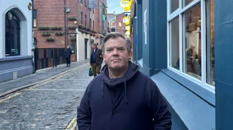 Paul is standing on hill street. He is wearing a navy jacket, and has short grey hair. Behind him is the cobbled street and footpaths. There are people walking behind him. There are red brick buildings and blue buildings in the background.