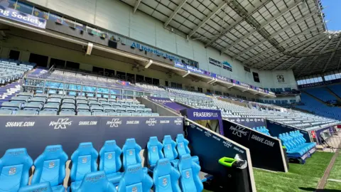 Blue players seats next to the player's entrance, seen from pitch side at a football stadium. Beyond, premier and hospitality zones can be seen.