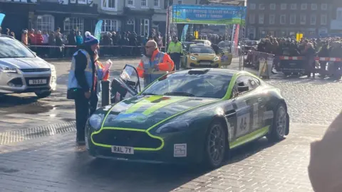 A black car with green stripes and race stickers is parked in the middle of a market square. Crowds stand behind red and white cordons, and there is a starting post in the background. Two officials, wearing hi-vis coats, are standing on the driver's side of the car, with the door open.