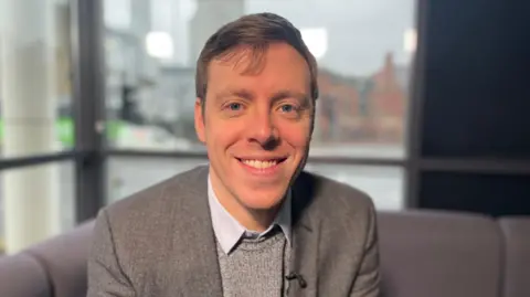 Mr Naish is wearing a grey jumper and jacket