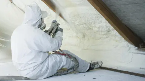 Getty Images Roof technician spraying foam insulation using component gun while wearing a white boilersuit