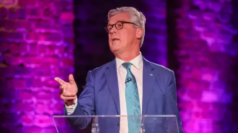 A man in a blue suit and teal tie speaks at a lectern, gesturing with his hand, against a purple-lit brick backdrop.