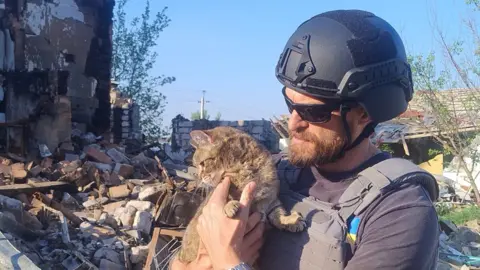 Lachlan Campbell holding a tabby cat. He is wearing a helmet and body armour among large piles of rubble. He has a brown beard and is wearing sunglasses.