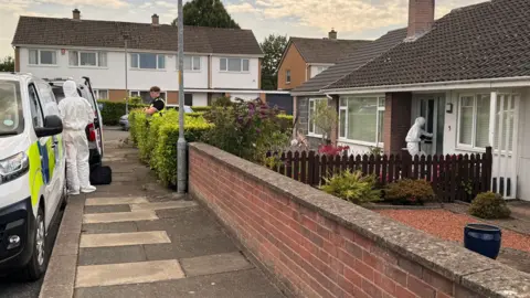 A bungalow on the right with a person in a white hazmat suit walking through the front door. There is a male police officer standing at the end of the garden path with brown hair and his arms crossed across his front. He is wearing a black top underneath a yellow hi-vis vest. There is another person in a white hazmat suit standing on the path outside the property with his back to the camera. They are standing between two police vans which have blue and yellow squares on the site. There are semi-detached two-storey houses on the street adjacent to the one with the bungalow.