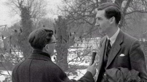 Black and white image of BBC reporter on left holding a microphone in front of the local doctor.  In the back ground snow can be seen between the trees.
