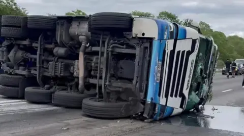 A blue and white lorry lying on it's side on the motorway carriagewat with it's wheels on show and it's windscreen smashed. a pool of diesel can be seen on the ground.