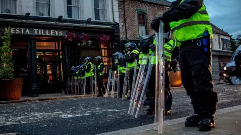 Police officers in riot gear stand in a line in the middle of a road in Sunderland. A pub is on the left, with a sign saying 'Vesta Tilleys'.