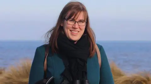Dr Teresa Frost Dr Teresa Frost frost smiling for the camera while outside near a body of water and ferns in the background. She is wearing glasses, a black scarf and a backpack. She also has a set of binoculars around her neck.