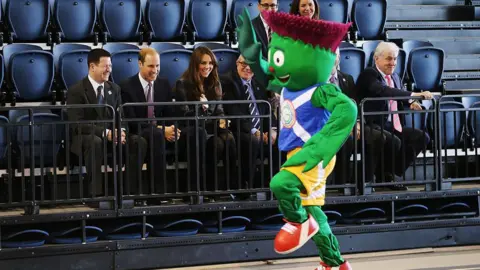Getty Images Clyde the green and purple animated thistle running past a stand of chair, filled with people inluding William and Kate who are laughing.