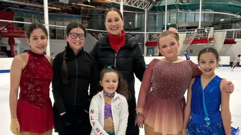 Rhiannon Scott A group of several skaters standing on  a rink. They are all children or teenagers, except for Rhiannon Scott, a woman with dark hair and wearing a black jacket with red top. She is standing in the centre of the group. 
