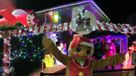 a person in a gingerbread man costume waving at the front of a house. Behind them is a merry christmas welcome sign covered in lights, and then festive decorations behind them. Over their shoulder is someone in a toy soldier costume.