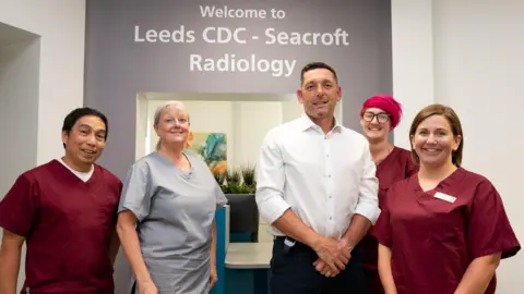 Five members of the hospital staff stand for a photo outside the diagnostic centre where the expansion is happening. Three nurses wear burgundy-coloured scrubs, one wears a grey scrub whilst a man wears a white shirt. 