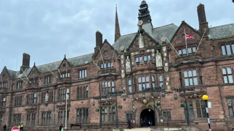 The exterior of Coventry City Council showing a brown brick building with arched windows and flagpoles.