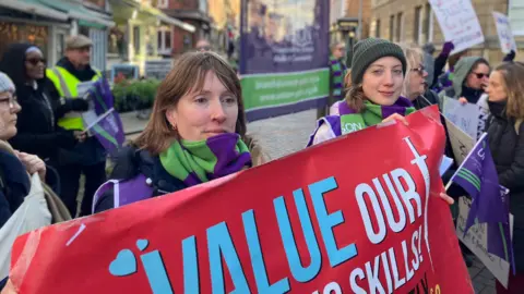 A woman with light brown hair to her shoulders holds up a red banner at a protest, along with another woman wearing a beanie. They are bundled up for the cold weather in green and purple scarves. Other people at the rally can be seen around them, holding flags and placards on a city centre, pedestrianised street.