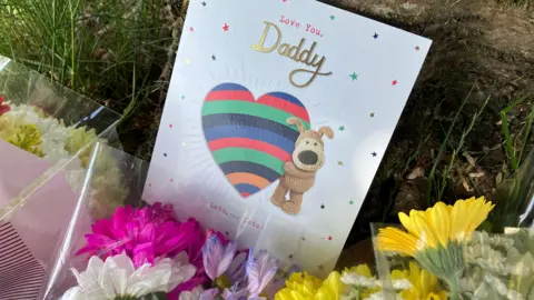 Mairead Smyth/BBC A card from a child with the words love you daddy rests between two bouquets of white, yellow and violet flowers