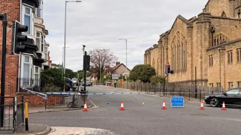 A police cordon near the junction between Kayll Road and Chester Road in Sunderland. The cordon, which is enforced with police tape and a blue police sign, shuts off a street with homes on the left side and a church on the right. To the left, part of a car can be seen in the bushes of a house. The railings near it are bent, implying it has smashed through them and into the house.
